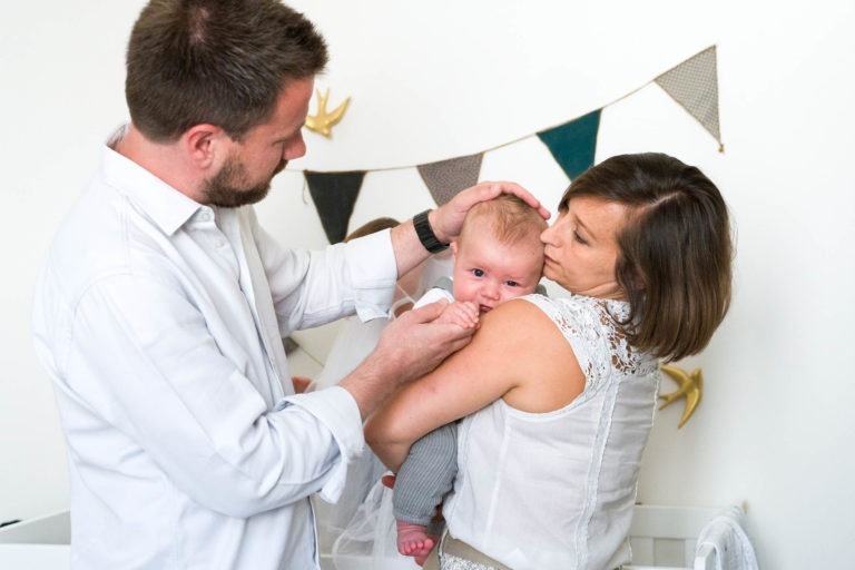 Un homme en chemise blanche touche doucement la tête d'un bébé tandis qu'une femme, également en tenue blanche, tient le bébé sur son épaule. Le bébé regarde directement l'objectif. Ils se trouvent dans une pièce au mur blanc décoré de banderoles triangulaires et de décorations murales, parfaites pour être immortalisées par n'importe quel photographe de famille Bordeaux.