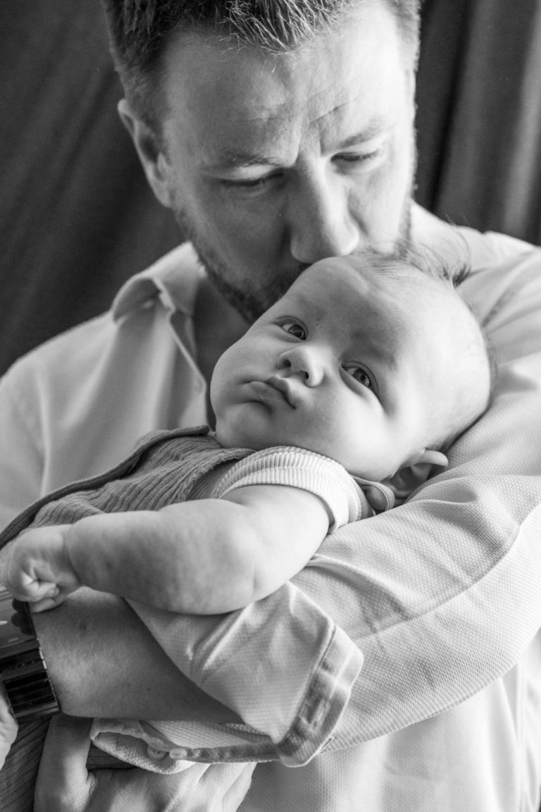 Un homme aux cheveux courts et à la barbe berce doucement un bébé dans ses bras. L'homme porte une chemise à manches longues et embrasse le bébé sur la tête. L'image, prise par le photographe de famille Bordeaux, montre le bébé dans une tenue à manches courtes regardant calmement au loin en noir et blanc.