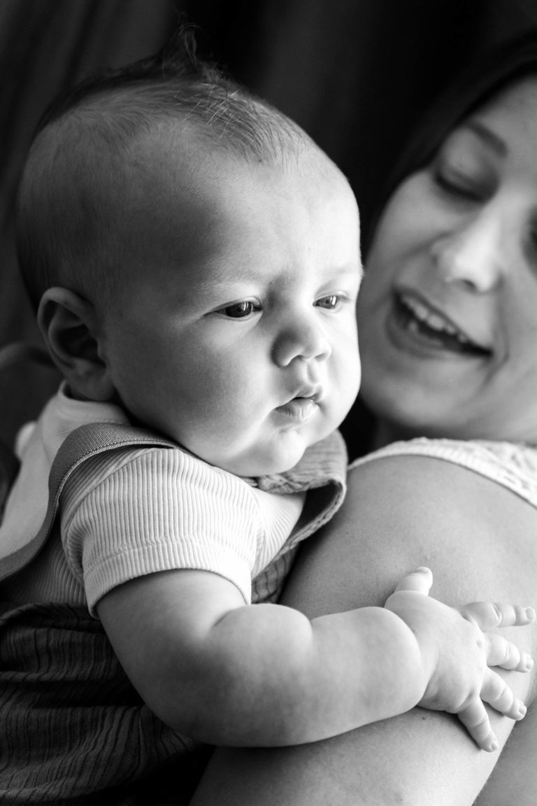 Une photo en noir et blanc prise par un photographe de famille de Bordeaux montre un bébé porté par-dessus l’épaule d’un adulte. Le bébé a les joues rebondies et porte un t-shirt sans manches. L’adulte, partiellement visible, a les cheveux longs et sourit joyeusement au bébé, qui regarde pensivement au loin.