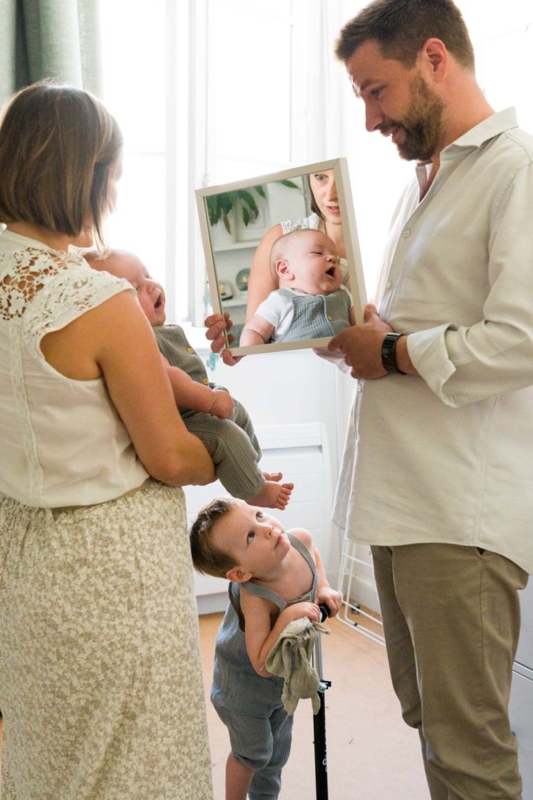 Une femme tient un bébé dans ses bras tandis qu'un homme lui montre son reflet dans un miroir. Un jeune enfant se tient à proximité, regardant le bébé avec curiosité. La scène, capturée par le photographe de famille Bordeaux, se déroule dans une pièce lumineuse, créant un moment familial chaleureux et intime.