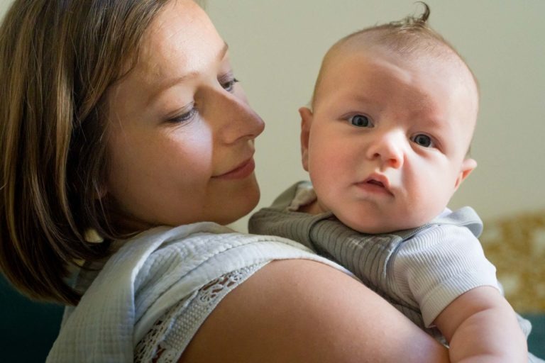 Une image en gros plan montre une femme tenant un bébé dans ses bras. La femme, aux cheveux châtains mi-longs et vêtue d'un vêtement blanc, regarde affectueusement le bébé. Le bébé, vêtu d'une tenue grise, a les cheveux clairs et regarde directement l'objectif avec une expression curieuse. Le photographe de famille Bordeaux capture parfaitement ce moment de tendresse.