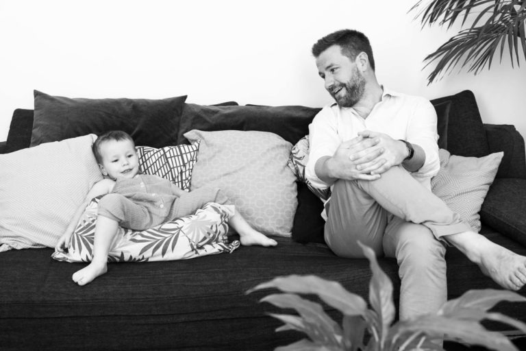 Une photo en niveaux de gris du photographe de famille Bordeaux montre un homme adulte assis sur un canapé, tourné vers un jeune enfant. L'homme sourit et se penche en avant, les mains jointes. L'enfant est allongé sur le même canapé, entouré de coussins. Tous deux semblent détendus et heureux. Une plante au premier plan ajoute une touche de nature.