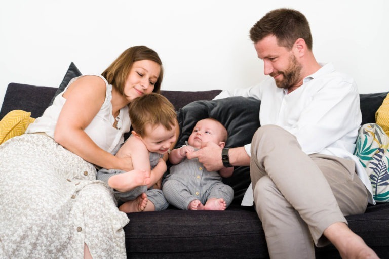 Une famille de quatre personnes est assise sur un canapé sombre. La mère, vêtue d’un haut blanc sans manches et d’une jupe à motifs, serre dans ses bras un jeune enfant. Le jeune enfant, habillé en gris, se penche vers un bébé tout gris tenu par le père, qui porte une chemise blanche et un pantalon beige. Ils sourient tous et interagissent chaleureusement – un moment idéal capturé par le photographe de famille Bordeaux.