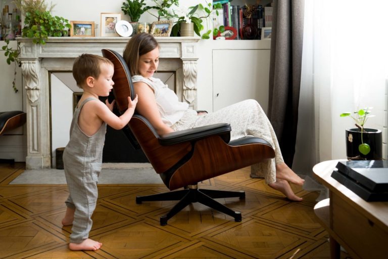 Un jeune enfant en salopette grise pousse une femme assise sur une chaise longue en bois dans un salon lumineux. La femme, vêtue d'une robe fleurie légère, sourit à l'enfant. Derrière eux se trouve un manteau blanc orné de photos encadrées, de plantes en pot et de livres. La lumière du soleil filtre à travers un rideau à droite, parfaitement capturée par notre photographe de famille Bordeaux.