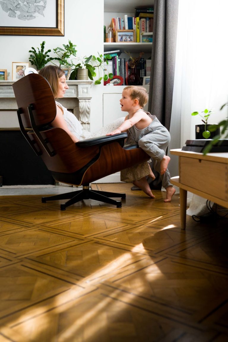 Une femme assise dans un fauteuil inclinable sourit à un jeune enfant qui grimpe sur l'accoudoir du fauteuil. La pièce est bien éclairée par la lumière naturelle, avec un plancher en bois, une cheminée, des étagères avec des livres et diverses plantes en pot. L'atmosphère est chaleureuse et familiale, idéale pour Photographe de famille Bordeaux.