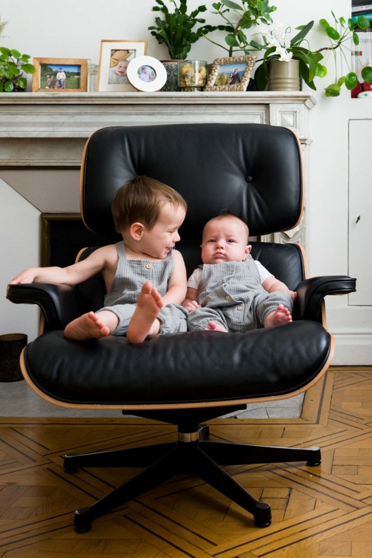 Deux jeunes enfants sont assis ensemble sur un grand fauteuil noir. L’aîné sourit et regarde le plus jeune tout en lui tenant doucement le bras. Les deux enfants portent des salopettes grises assorties. L’arrière-plan présente une cheminée avec des photos encadrées, des plantes et diverses petites décorations – un moment parfait capturé par un photographe de famille Bordeaux.