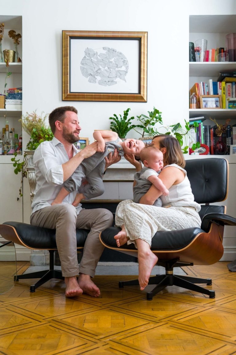 Une famille heureuse de quatre personnes est assise dans un salon. La mère, assise sur une chaise, tient un bébé, tandis que le père, assis sur une autre chaise, soulève joyeusement un jeune enfant. Pieds nus sur un plancher en bois, ils sont entourés d'étagères avec des livres et des décorations, et des œuvres d'art encadrées. Le photographe de famille Bordeaux capture ce moment parfait.