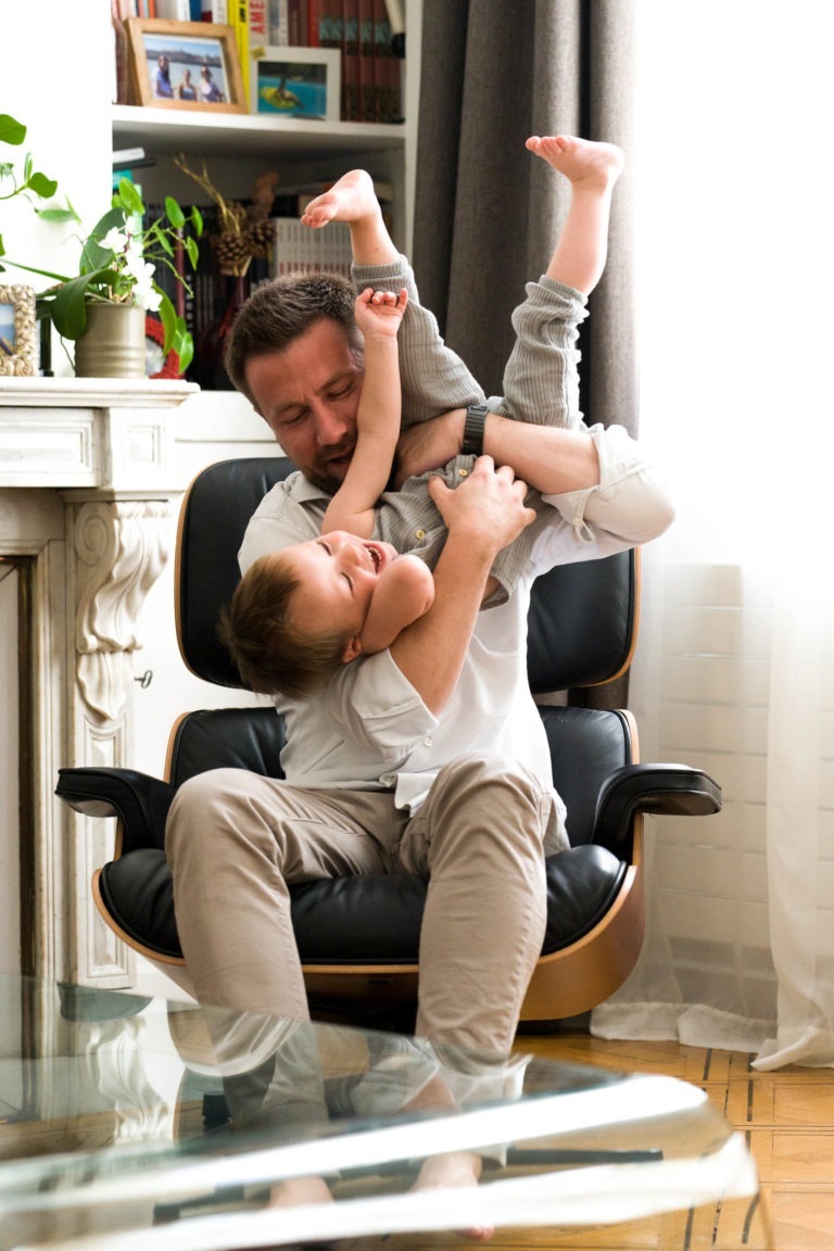 Un homme assis sur un fauteuil noir moderne aux accents de bois tient de manière ludique un jeune enfant la tête en bas. L'enfant, vêtu d'un pantalon gris clair, rit les pieds en l'air. Captured par le photographe de famille Bordeaux, la pièce confortable comprend des étagères, des plantes et une cheminée chaleureuse en arrière-plan.