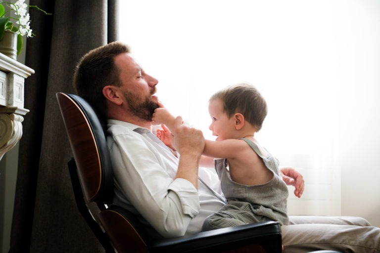 Un homme aux cheveux noirs courts et à la barbe, vêtu d'une chemise blanche, est assis sur une chaise noire et en bois moderne. Un jeune enfant en salopette grise est assis sur ses genoux, touchant joyeusement le visage de l'homme. Tous deux semblent heureux. Une fenêtre avec des rideaux blancs transparents laissant entrer une lumière vive se trouve à l'arrière-plan - la scène parfaite pour tout photographe de famille Bordeaux.