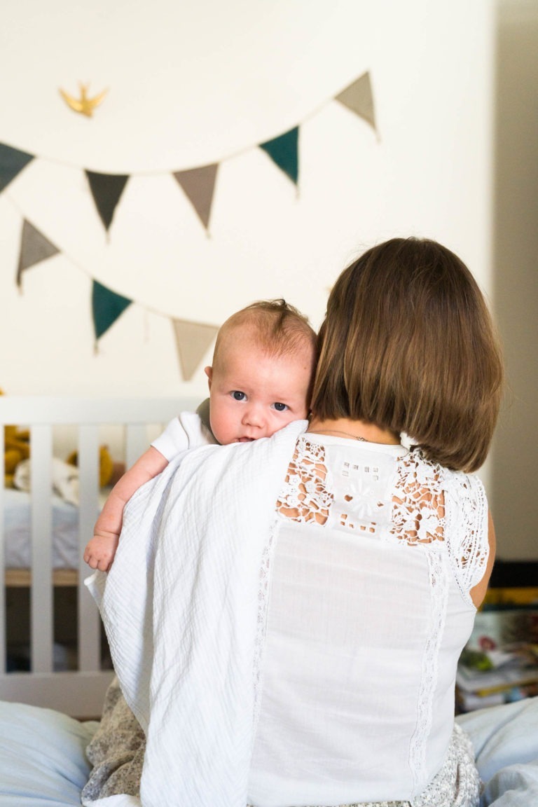 Une femme aux cheveux bruns courts est vue de dos, tenant un bébé sur son épaule. Le bébé, vêtu d'une tenue blanche, regarde directement l'objectif. La femme porte un haut blanc crocheté. En arrière-plan, un berceau blanc et des banderoles triangulaires colorées ajoutent du charme à ce moment parfait de Photographe de famille Bordeaux.