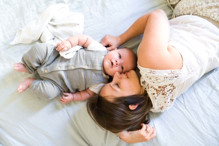 Une femme est allongée sur le dos dans son lit à côté d'un bébé qui porte une combinaison rayée grise avec des boutons. La femme, vêtue d'un haut en dentelle blanche, regarde avec amour le bébé qui est partiellement recouvert d'une couverture légère. Tous deux sont sur un couvre-lit bleu clair, capturant un moment parfait pour tout photographe de famille bordelais.