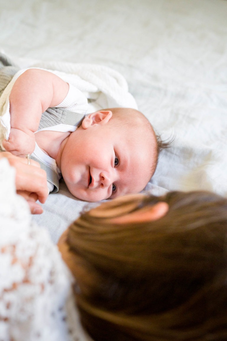 Un bébé, allongé sur une surface douce, lève les yeux et sourit à une personne aux cheveux bruns qui se trouve au premier plan. Le bébé est enveloppé dans une couverture blanche et porte un haut de couleur claire. L'atmosphère semble tendre et intime, parfaitement capturée par le photographe de famille Bordeaux.