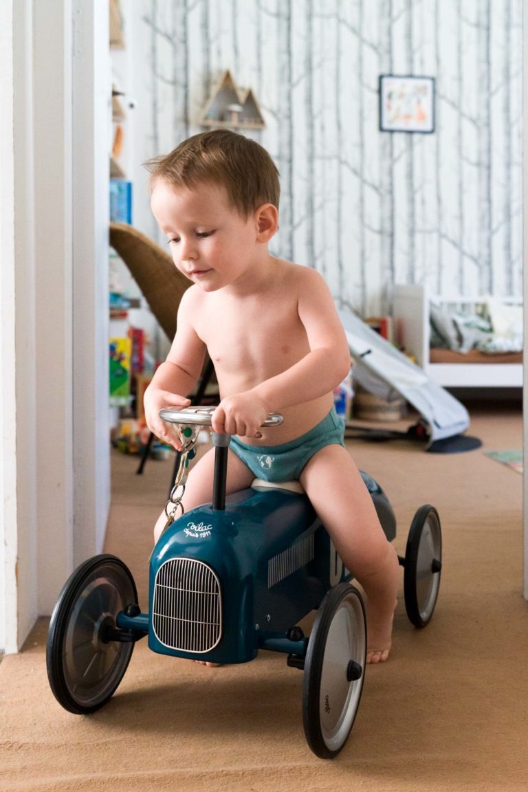 Un jeune enfant en sous-vêtements bleus conduit une petite voiture à pédales vert foncé à l'intérieur. La pièce est décorée d'un papier peint à motifs d'arbres, d'un tapis marron et de divers jouets et livres en arrière-plan. Photographié par un photographe de famille Bordeaux, l'enfant aux cheveux bruns courts et aux pieds nus est concentré sur la conduite de la voiture.