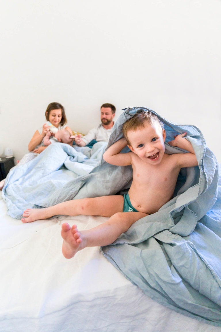 Un enfant heureux aux cheveux ébouriffés joue joyeusement sous une couverture au pied d'un lit. En arrière-plan, un homme et une femme sont assis sur le lit, souriant et s'occupant d'un bébé dans leurs bras. Capturée par le photographe de famille Bordeaux, la scène respire la chaleur et la convivialité familiale dans une chambre à coucher décontractée et confortable.