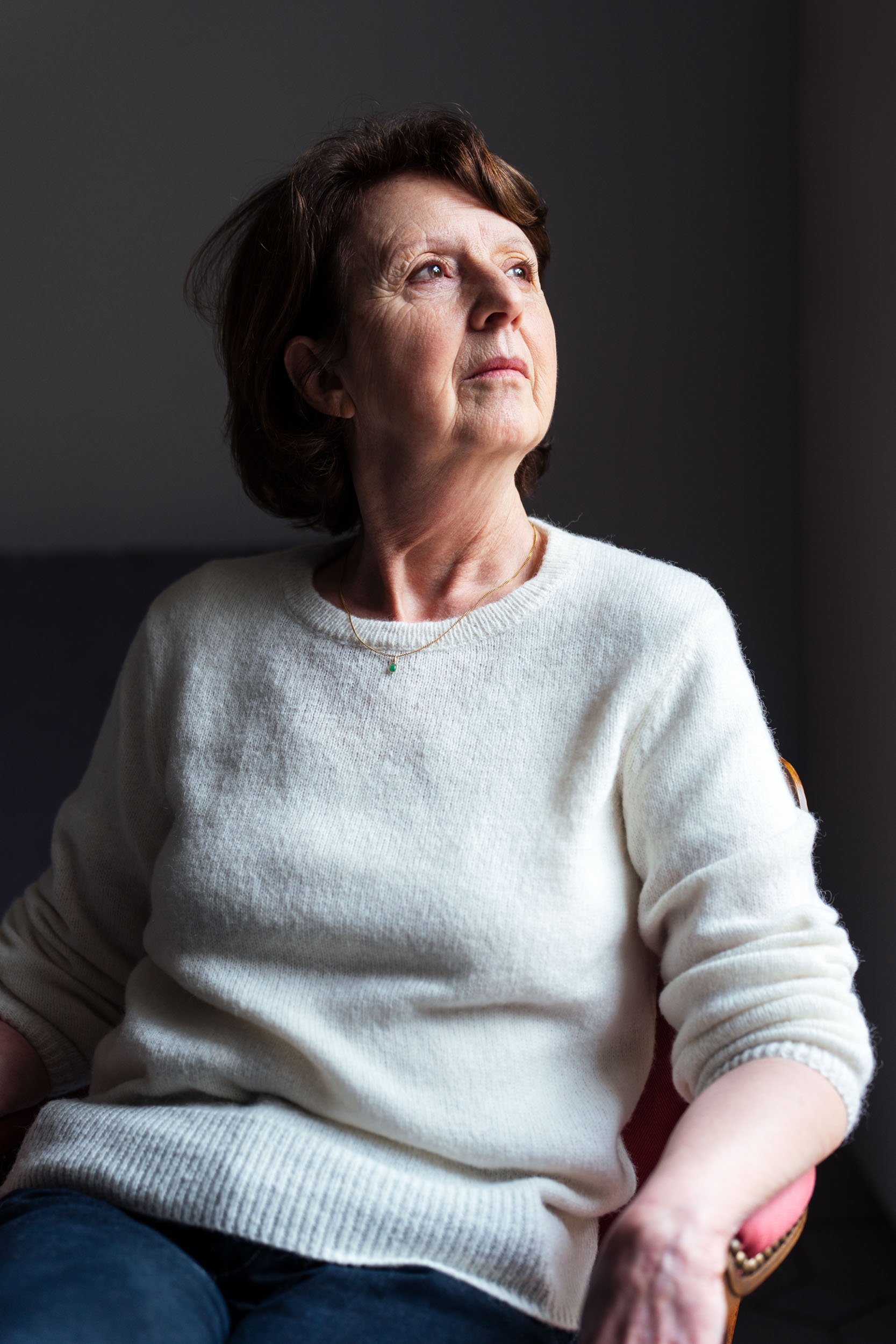 Une femme aux cheveux bruns courts est assise sur un fond sombre, vêtue d'un pull blanc et d'un jean bleu. Elle regarde vers sa droite avec une expression pensive, et la lumière du jour illumine le côté gauche de son visage et de son corps, créant un contraste avec l'environnement plus sombre derrière elle - un exemple de portraits professionnels à Bordeaux.