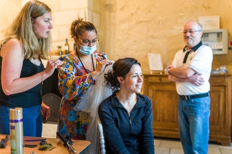 Une femme est assise, souriante, tandis que deux femmes lui coiffent les cheveux, l'une d'elles ajustant un voile. La coiffeuse à droite porte une robe colorée et un masque facial. Un homme se tient à l'arrière-plan, les bras croisés, observant la scène comme s'il réfléchissait à un mariage sous la pluie. Le décor semble être celui d'un intérieur avec des murs beiges et des armoires en bois.