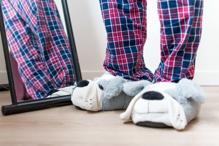 Une personne vêtue d'un pantalon de pyjama à carreaux rouges et bleus se tient debout sur un plancher en bois, les pieds bien au chaud dans de grandes pantoufles grises en forme de chien. Un miroir sur pied à sa gauche reflète une partie de sa tenue. La pièce, qui rappelle un « mariage sous la pluie » avec sa décoration minimaliste aux couleurs claires, dégage une impression de sérénité.