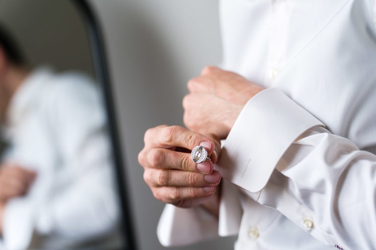 Une personne vêtue d'une chemise blanche ajuste un bouton de manchette en argent à son poignet, peut-être pour un mariage sous la pluie. Le reflet de son torse est visible dans un miroir derrière elle. Le décor semble se dérouler à l'intérieur, peut-être en prévision de l'événement officiel. L'accent est mis sur les mains et le bouton de manchette.