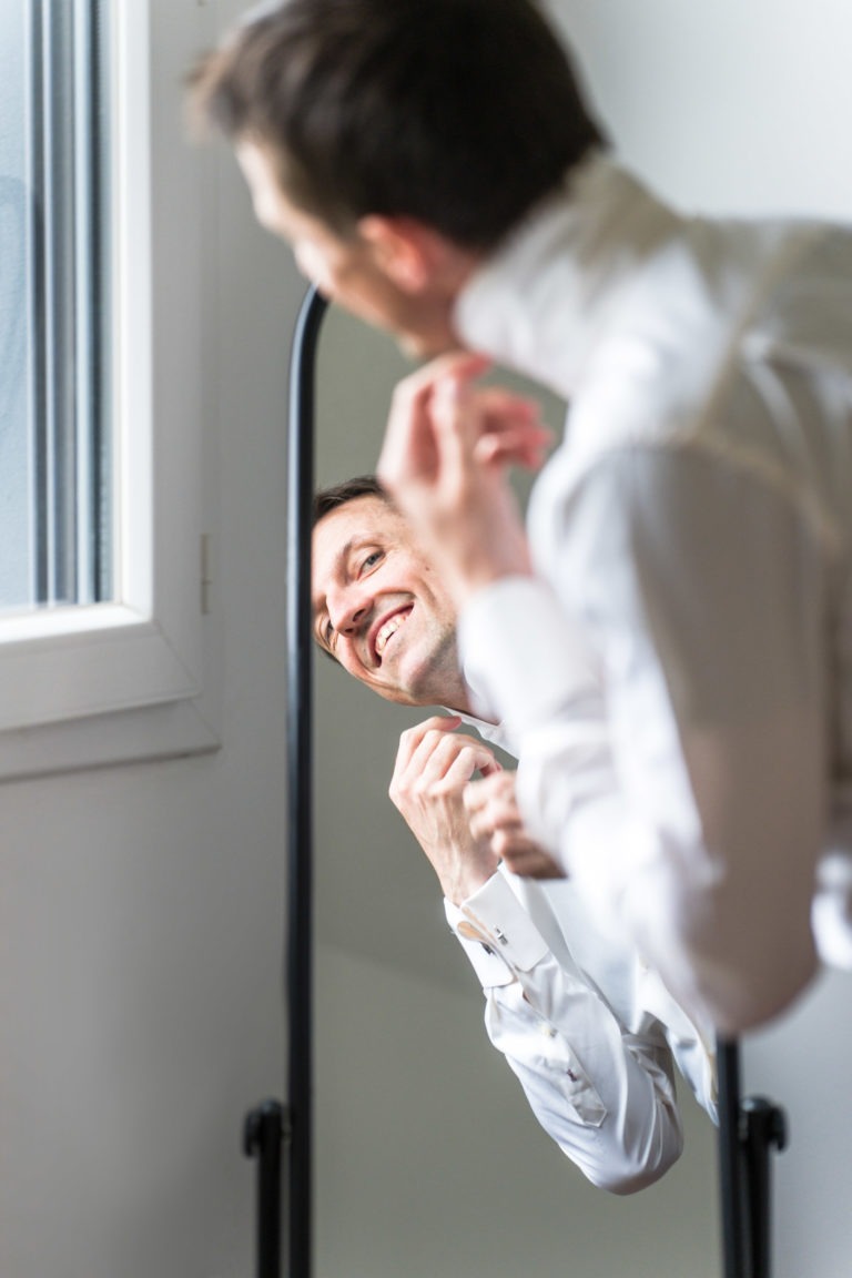 Un homme se tient devant un miroir, boutonnant le col de sa chemise blanche. Il sourit et incline légèrement la tête, tandis que la lumière du soleil entre par une fenêtre voisine, rappelant une scène de « Mariage sous la pluie ». Le reflet montre son visage et le haut de son corps alors qu'il termine sa tenue.