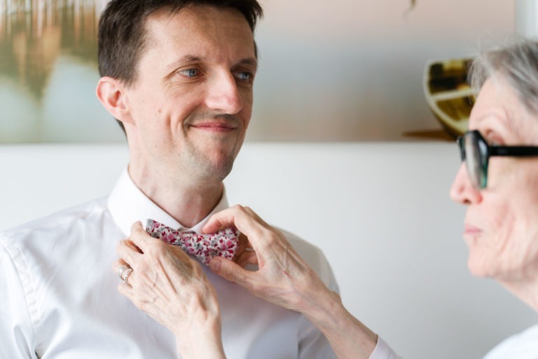 Un homme portant une chemise blanche sourit tandis qu'une femme plus âgée l'aide à ajuster son nœud papillon fleuri, se préparant pour un mariage sous la pluie. La femme, aux cheveux gris courts et portant des lunettes, se concentre sur le nœud papillon avec ses deux mains. L'arrière-plan est légèrement flou, avec une œuvre d'art murale encadrée.