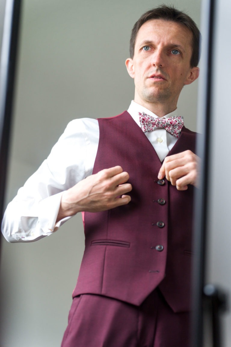 Un homme aux cheveux courts ajuste les boutons de son gilet bordeaux devant un miroir, se préparant peut-être pour un mariage sous la pluie. Il porte une chemise blanche et un nœud papillon fleuri. L'arrière-plan est neutre, mettant l'accent sur sa tenue formelle et sa préparation.