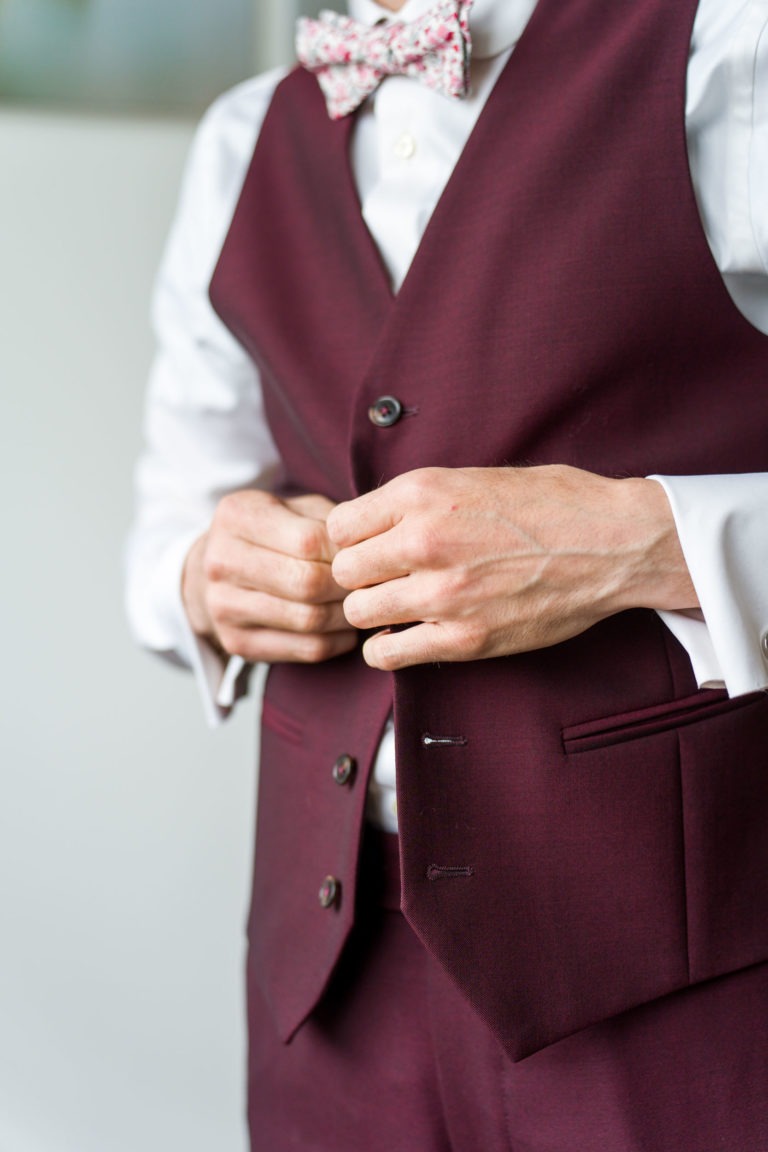 Une personne portant un gilet marron sur une chemise blanche boutonne son gilet, se préparant pour un mariage sous la pluie. L'image est centrée sur le torse et les mains, sans tête ni détails d'arrière-plan visibles. Le gilet a trois boutons, dont deux boutonnés, et deux poches avant.