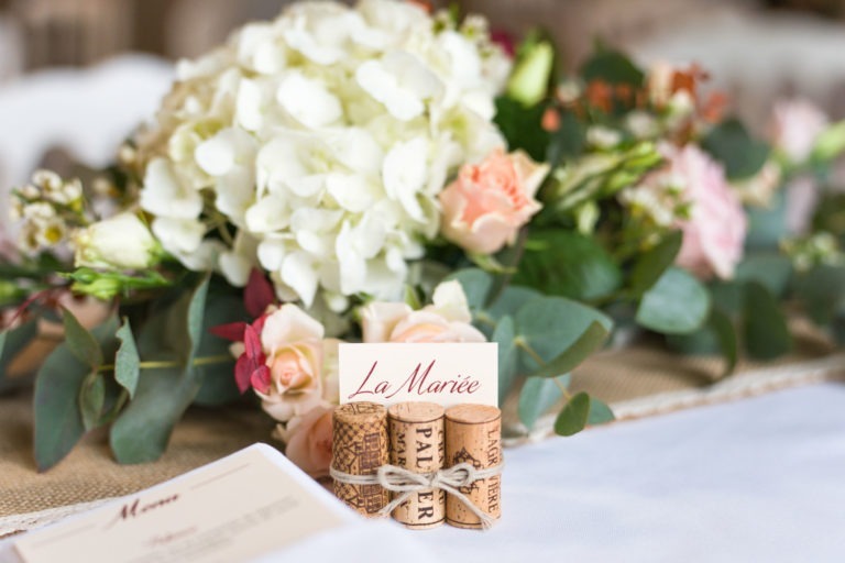 Gros plan d'un centre de table de mariage avec un bouquet d'hortensias blancs, de roses roses, de verdure et de petites fleurs. À l'avant, des bouchons de vin personnalisés attachés avec de la ficelle maintiennent une carte sur laquelle est écrit « La Mariée ». La mise en scène évoque le charme d'un *mariage sous la pluie*. Une carte de menu est partiellement visible au premier plan.