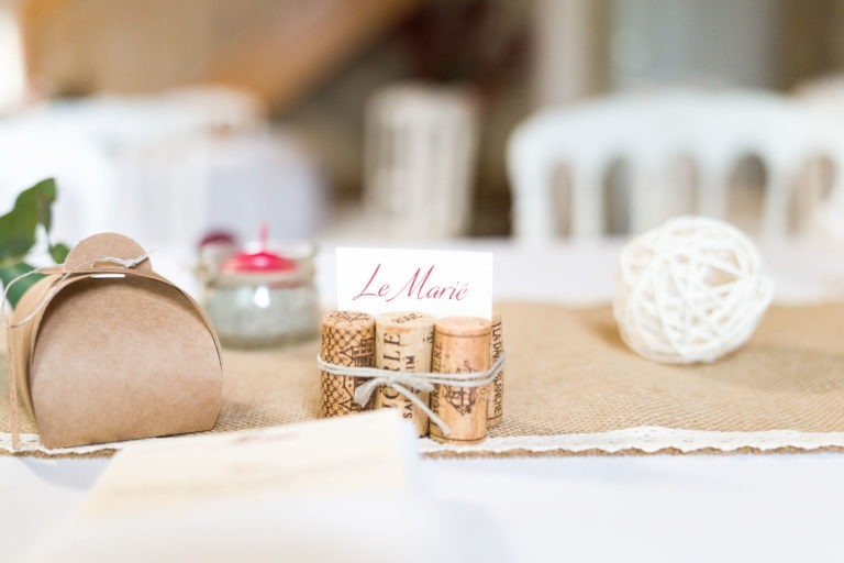 Une décoration de table composée d'un marque-place étiqueté "Le Marié" maintenu par des bouchons de vin, d'une petite boîte cadeau marron, d'une bougie allumée dans un bocal et d'une boule en rotin blanc. Le tout est présenté sur un chemin de table en toile de jute et dentelle avec des chaises blanches floues en arrière-plan, évoquant le charme de "Mariage sous la pluie".