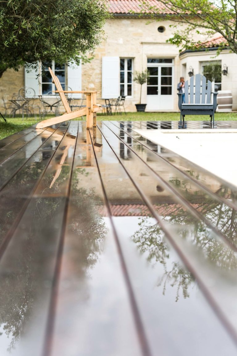 Au premier plan, une terrasse en bois moderne avec des reflets d'arbres et de chaises. À l'arrière-plan, une maison en pierre avec de grandes fenêtres, des volets blancs et un toit rougeâtre se dresse au milieu d'arbres verts et d'une pelouse. Des meubles d'extérieur sont dispersés autour, notamment des bancs et des chaises. Deux personnes sont visibles près de la maison, se remémorant peut-être leur « mariage sous la pluie ».