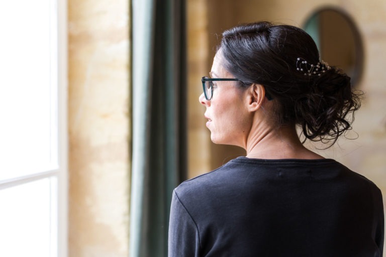 Une femme aux cheveux noirs tirés en arrière en un chignon lâche maintenu par une pince à cheveux se tient debout à l'intérieur, face à une fenêtre où la lumière naturelle illumine son visage. Elle porte des lunettes et un haut à manches longues foncé. Un rideau vert et un miroir rond sont visibles en arrière-plan, rappelant "Mariage sous la pluie".