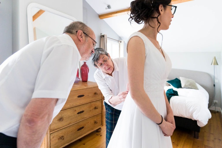 Dans une pièce lumineuse, deux personnes âgées assistent une jeune femme en robe de mariée. La femme, aux cheveux noirs relevés en chignon, reste immobile tandis que la femme âgée attache sa robe dans le dos. Un homme âgé observe attentivement, se remémorant leur propre « mariage sous la pluie ». La pièce est décorée d'une commode en bois, d'un miroir et d'un lit en arrière-plan.