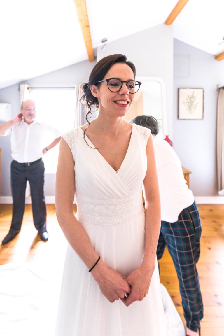 Une mariée souriante, vêtue d'une robe blanche sans manches avec des détails en dentelle, se tient dans une pièce lumineuse. Elle porte des lunettes noires et ses cheveux sont coiffés en chignon lâche. Deux hommes ajustent leur tenue derrière elle, se remémorant peut-être leur propre mariage sous la pluie. La pièce est dotée de poutres en bois, d'œuvres d'art encadrées sur les murs et d'un plancher en bois.