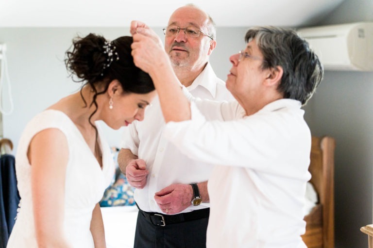 Une femme en robe blanche, vraisemblablement une mariée, s'incline légèrement tandis qu'une autre femme ajuste sa perruque. Un homme plus âgé se tient derrière elles et les observe attentivement. Elles se trouvent dans une pièce aux murs de couleur neutre, mettant en valeur un climatiseur et des parties de meubles en bois. On se croirait dans un moment serein avant un mariage sous la pluie.