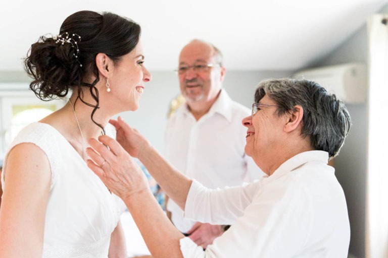 Une mariée souriante, les cheveux noirs relevés en chignon et ornés d'un postiche décoratif, se tient dans une pièce blanche. Elle est en train d'être ajustée par une femme plus âgée aux cheveux gris courts et aux lunettes, tandis qu'un homme plus âgé portant des lunettes et une barbe regarde, souriant lui aussi. Malgré la pluie qui fait penser à un « mariage sous la pluie », tout le monde semble joyeux et affectueux.