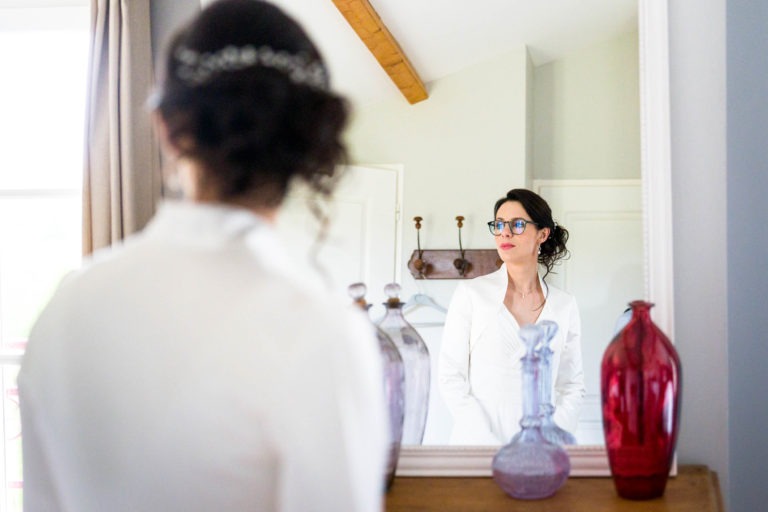 Une femme en robe blanche se tient devant un miroir et contemple son reflet. Elle a les cheveux noirs coiffés en boucles lâches et est ornée d'un délicat diadème. La pièce, qui rappelle "Mariage sous la pluie", présente des murs de couleur claire, un plafond à poutres en bois et des vases décoratifs en verre sur la commode en bois à côté du miroir.