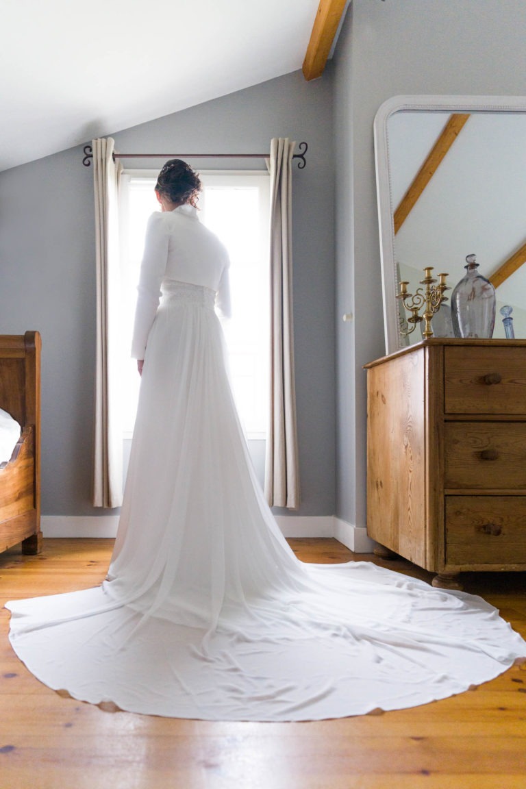 Une mariée vêtue d'une élégante robe blanche à longue traîne se tient debout face à une fenêtre dans une pièce confortable et lumineuse. Ses cheveux sont relevés et elle est entourée de meubles en bois rustiques, dont une commode avec des objets décoratifs et un lit partiellement visible à gauche, rappelant son charmant mariage sous la pluie.