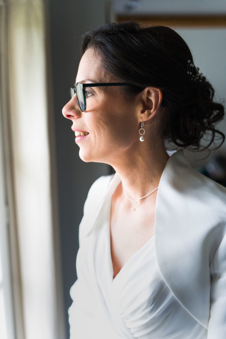 Une femme aux cheveux noirs attachés en arrière, portant des lunettes, regarde par la fenêtre. Vêtue d'une tenue blanche avec un décolleté en V profond et un collier délicat, elle incarne l'élégance. Ses boucles d'oreilles à pendentifs circulaires captent la douce lumière naturelle qui illumine son visage, comme si elle méditait sur un "Mariage sous la pluie".