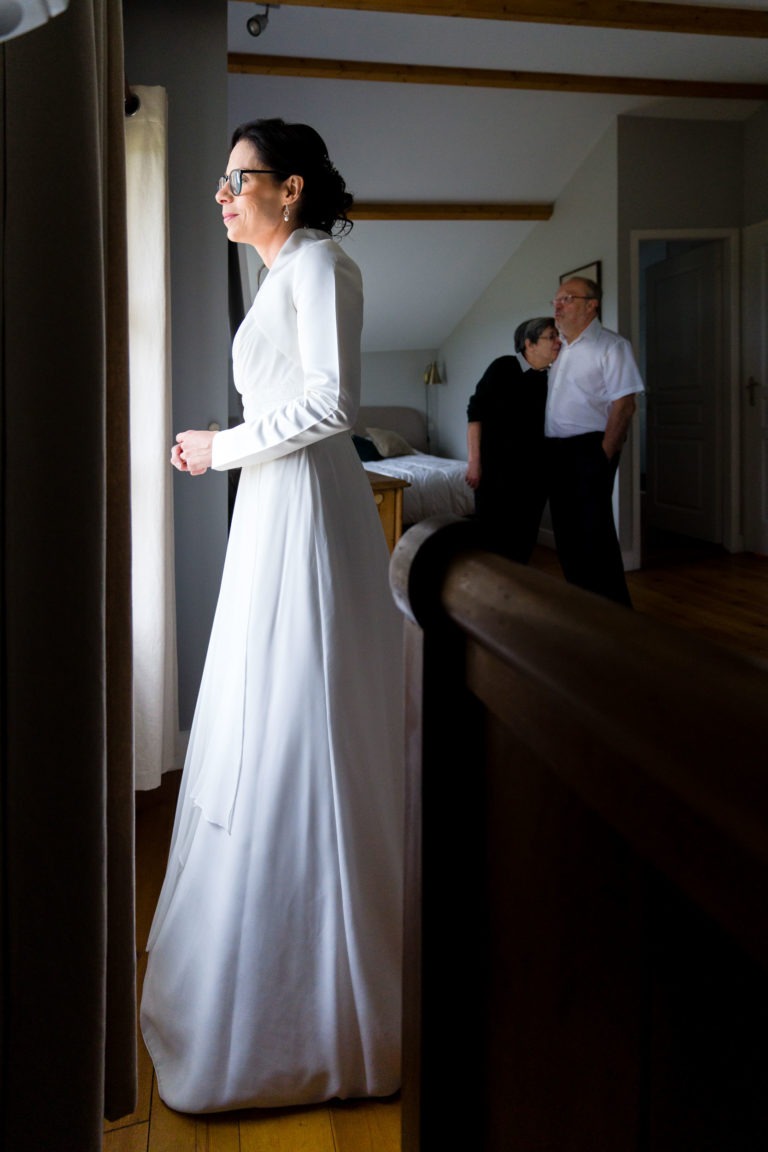 Une femme en robe blanche à manches longues se tient près d'une fenêtre, regardant dehors d'un air pensif. Ses cheveux sont bien coiffés et elle porte des lunettes. En arrière-plan, un couple, tous deux habillés de façon décontractée, se tient près du lit, engagé dans une conversation tranquille sur leur « Mariage sous la pluie ».