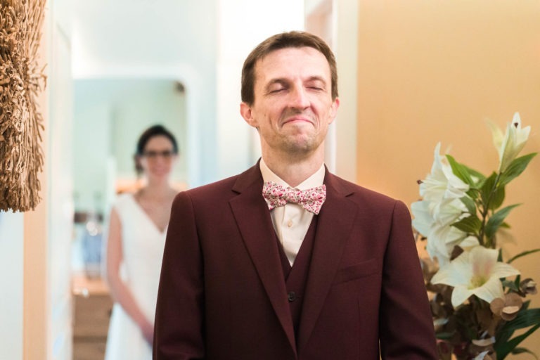 Un homme en costume marron avec un nœud papillon fleuri se tient debout, les yeux fermés et un sourire satisfait. Derrière lui, une femme en robe de mariée blanche est légèrement floue, semblant s'approcher de lui. Le décor semble avoir un éclairage intérieur chaleureux avec des plantes décorant un côté de la scène, évoquant le charme du « mariage sous la pluie ».