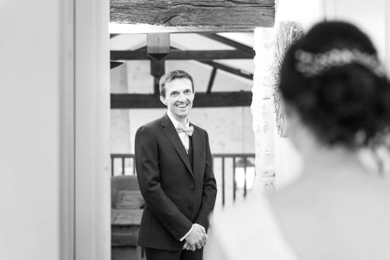 Un marié en costume sombre et nœud papillon sourit chaleureusement en regardant la mariée. L'espace intérieur rustique, avec ses poutres en bois apparentes et ses murs en pierre, évoque un « mariage sous la pluie » chaleureux. La mariée, partiellement visible de dos, porte une robe blanche ornée d'accessoires pour cheveux.