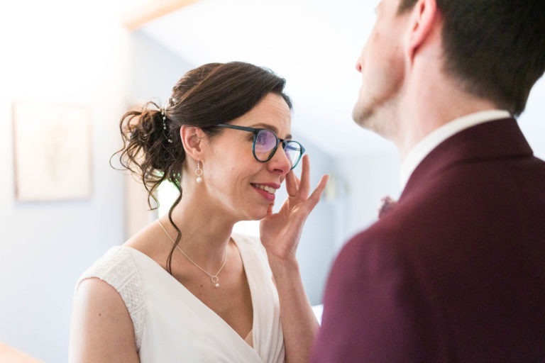 Une mariée, vêtue d'une robe blanche et de lunettes, essuie une larme de son visage avec sa main. Elle a les cheveux noirs relevés et regarde vers un homme en costume bordeaux, qui tourne le dos à la caméra. La pièce à la lumière tamisée présente des murs aux couleurs claires et des œuvres d'art encadrées, capturant l'essence du « mariage sous la pluie ».