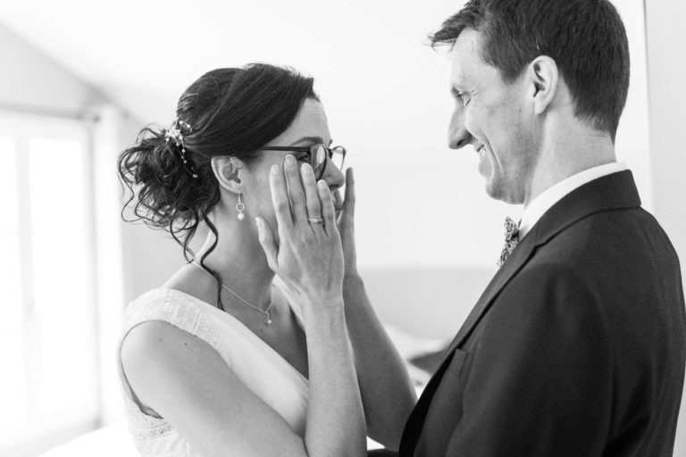 Un couple de mariés partage un moment intime le jour de leur mariage. Sous le thème du mariage sous la pluie, la mariée a les cheveux relevés en chignon élégant avec un accessoire décoratif et porte des lunettes, une robe blanche et des boucles d'oreilles. Le marié, en costume sombre et cravate, la regarde avec amour tandis qu'elle touche son visage, semblant ému et heureux.