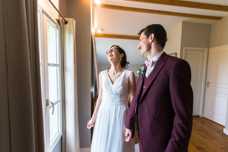 Un couple de mariés se tenant la main regardent par la fenêtre. La mariée, vêtue d'une robe blanche et de lunettes, sourit tandis que le marié, en costume bordeaux et nœud papillon, regarde vers la fenêtre. Malgré le mariage sous la pluie à l'extérieur, les poutres en bois et les murs clairs de la pièce ajoutent de la chaleur à la scène.