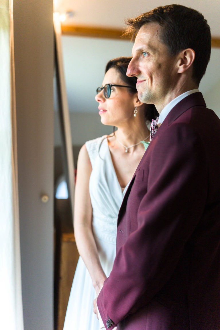 Un homme en costume marron et une femme en robe blanche se tiennent côte à côte, regardant par la fenêtre. La femme porte des lunettes et un collier, et l'homme a un nœud papillon. Ils semblent pensifs et satisfaits, avec une douce lumière du jour illuminant leurs visages, évoquant l'ambiance sereine de "Mariage sous la pluie".