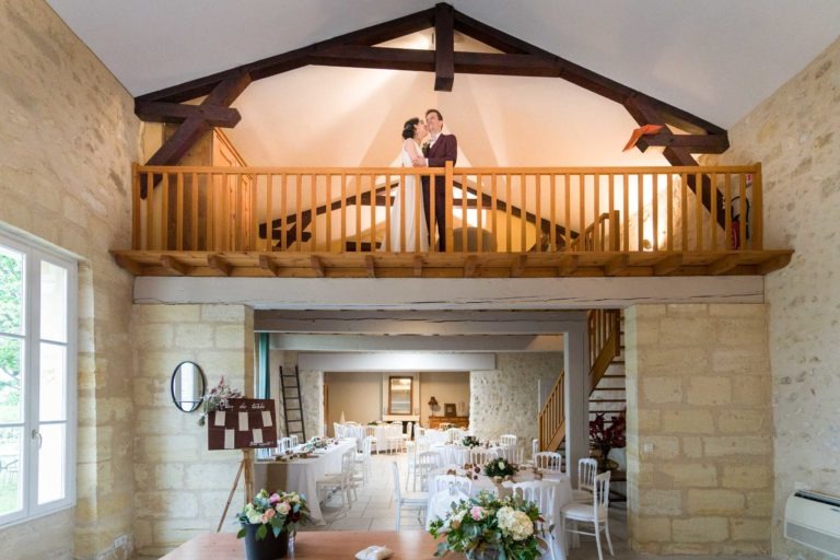 Un couple se tient sur un balcon en bois surplombant une spacieuse salle de réception de mariage dans un lieu rustique. L'espace de réception en contrebas comprend des tables rondes dressées pour un repas avec des chaises blanches et des centres de table floraux. Les poutres en bois et les murs en pierre ajoutent au charme rustique, avec une lumière naturelle remplissant la pièce, rappelant l'atmosphère de rêve d'un mariage sous la pluie.