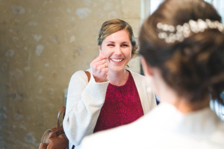 Une femme souriante, vêtue d'une robe en dentelle bordeaux et d'un cardigan beige, dessine un coeur avec sa main. Elle se tient debout devant une autre femme qui lui tourne le dos, dévoilant ses cheveux noirs ornés d'un postiche décoratif. Le fond évoque le charme de "Mariage sous la pluie" avec son mur doux, beige et texturé.