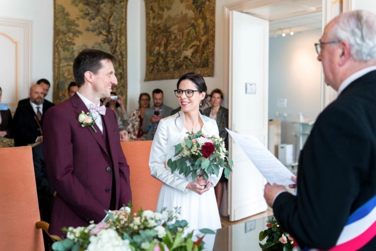 Un couple se tient face à face, sur le point de se marier lors d'une journée mémorable de mariage sous la pluie. Le marié, en costume bordeaux et nœud papillon, sourit à la mariée dans sa robe blanche et ses lunettes, tenant un bouquet de fleurs rouges et blanches. L'officiant, en costume sombre avec une ceinture tricolore, lit un document pendant que les invités regardent en arrière-plan.