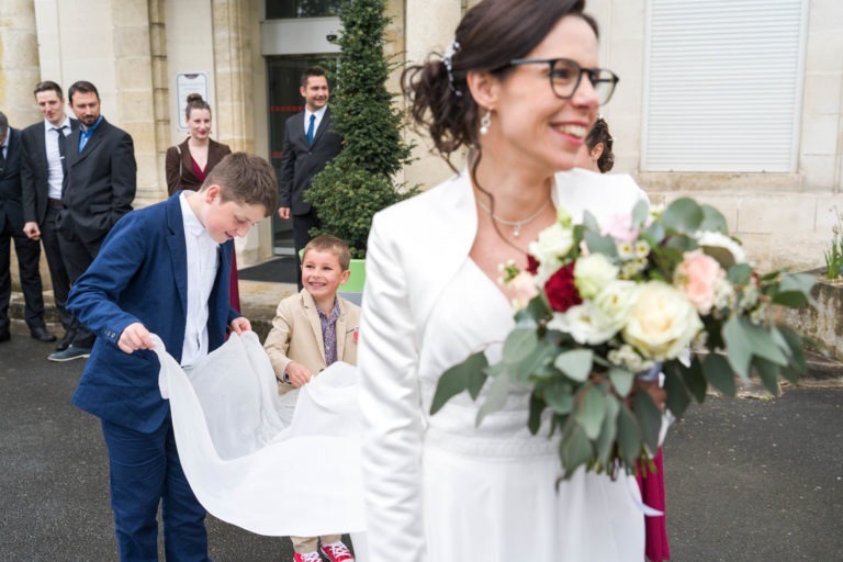 Une mariée souriante, vêtue d'une robe blanche et de lunettes, tient un bouquet de fleurs. Deux garçons en costume, l'un bleu et l'autre en baskets beiges et rouges, rient en soulevant la traîne de sa robe. En arrière-plan, des invités bien habillés se tiennent près d'un bâtiment en pierre. La scène animée embrasse l'esprit d'un "Mariage sous la pluie".