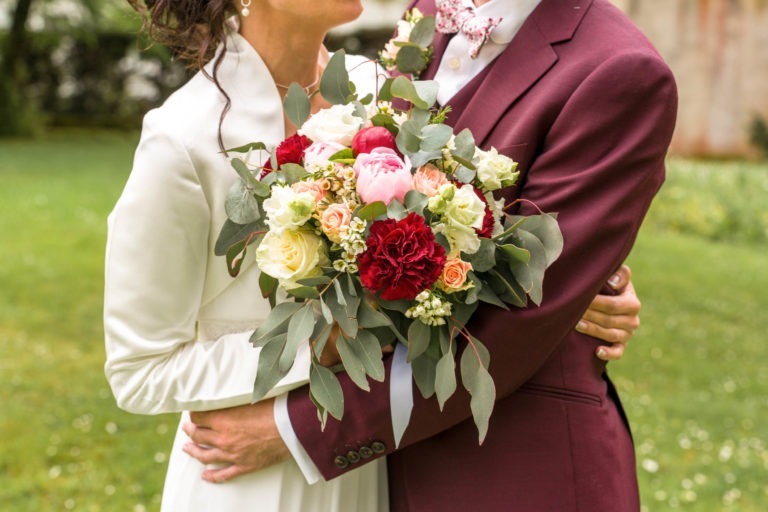 Un couple en tenue de mariage s'embrasse à l'extérieur. La mariée, en robe blanche, tient un bouquet de roses et un assortiment de fleurs rouges, roses et blanches avec des feuilles d'eucalyptus. Le marié, en costume bordeaux et nœud papillon fleuri, a un bras autour de sa taille. Ils ont l'air radieux malgré le fait que ce soit un mariage sous la pluie.