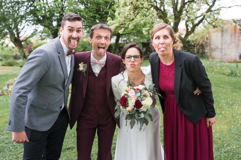 Une scène de mariage avec quatre personnes posant à l'extérieur, faisant des grimaces malgré la bruine. Le marié, vêtu d'un costume marron et d'un nœud papillon, se tient à côté de la mariée en robe blanche tenant un bouquet. Ils sont flanqués de deux invités, l'un en costume gris et l'autre en robe rouge avec un blazer noir. Un mariage sous la pluie peut être délicieux !